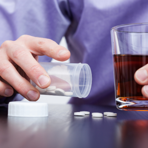 Man with alcoholic drink in one hand and pouring pills onto a table with other hand. Photo shows hands only.