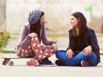 Two young adult women talking, with one sitting on a skateboard and the other sitting on the ground cross-legged.