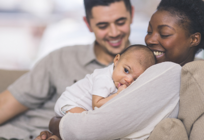 Woman Holding Baby, Smiling; Man in Background Looking at Baby and Smiling