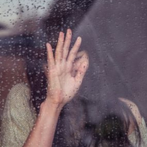 Person standing in front of window on rainy day with hand in front of their face.