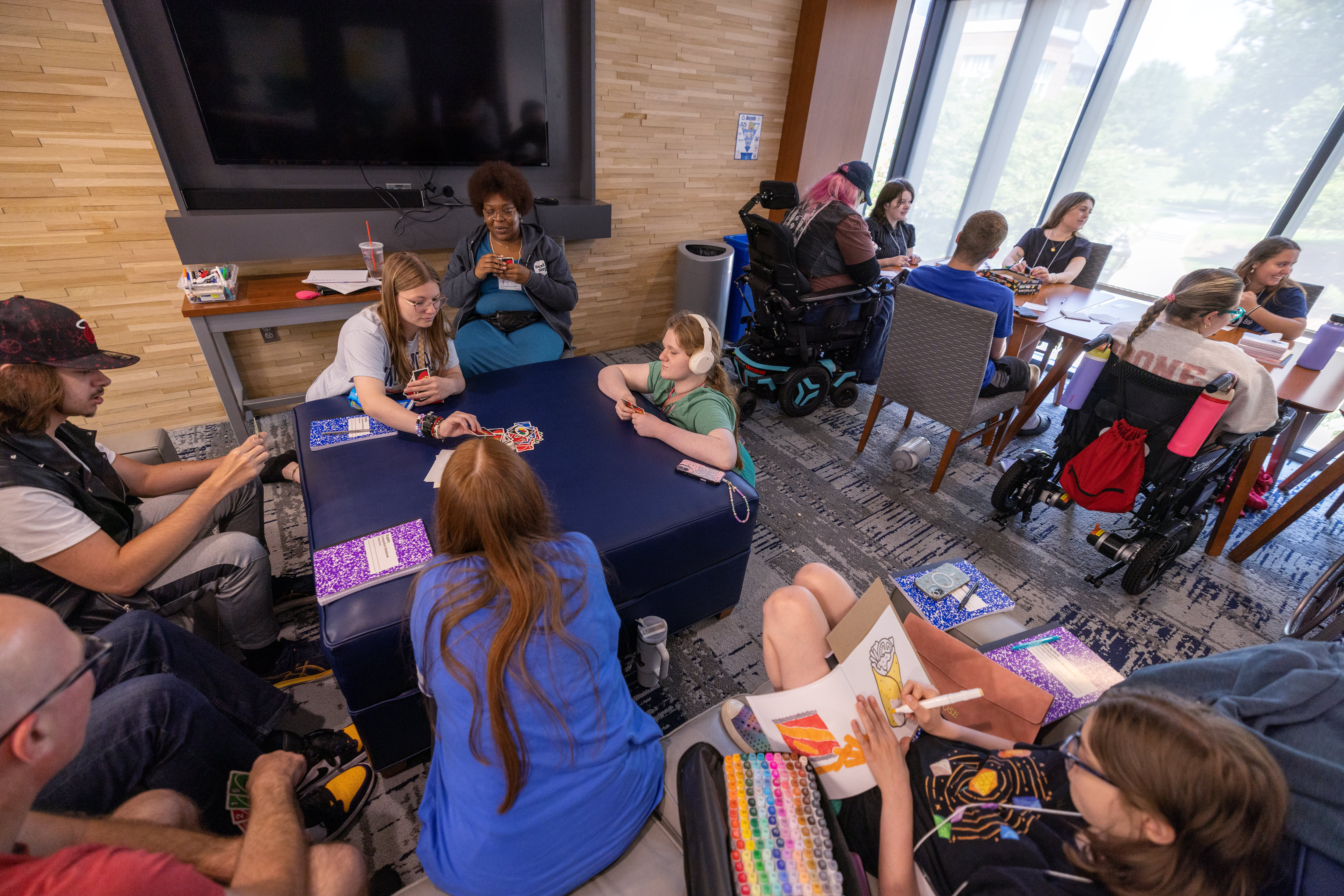 Young adults gather around a table to play a game. Other young adults sitting in different parts of the room engaged in different activities.