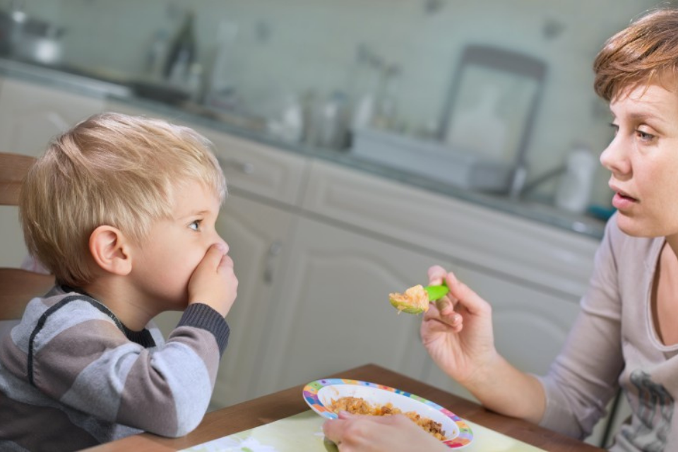 Woman trying to feed young child. Child is covering mouth with hands.