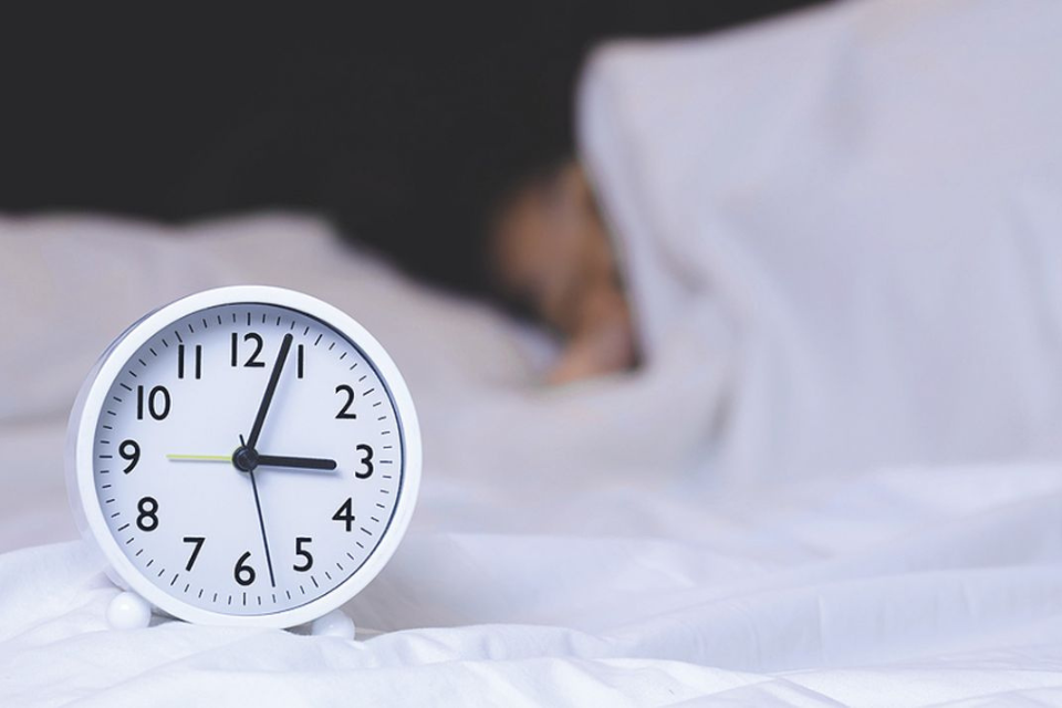 Alarm clock in foreground, person curled up under white covers in background.