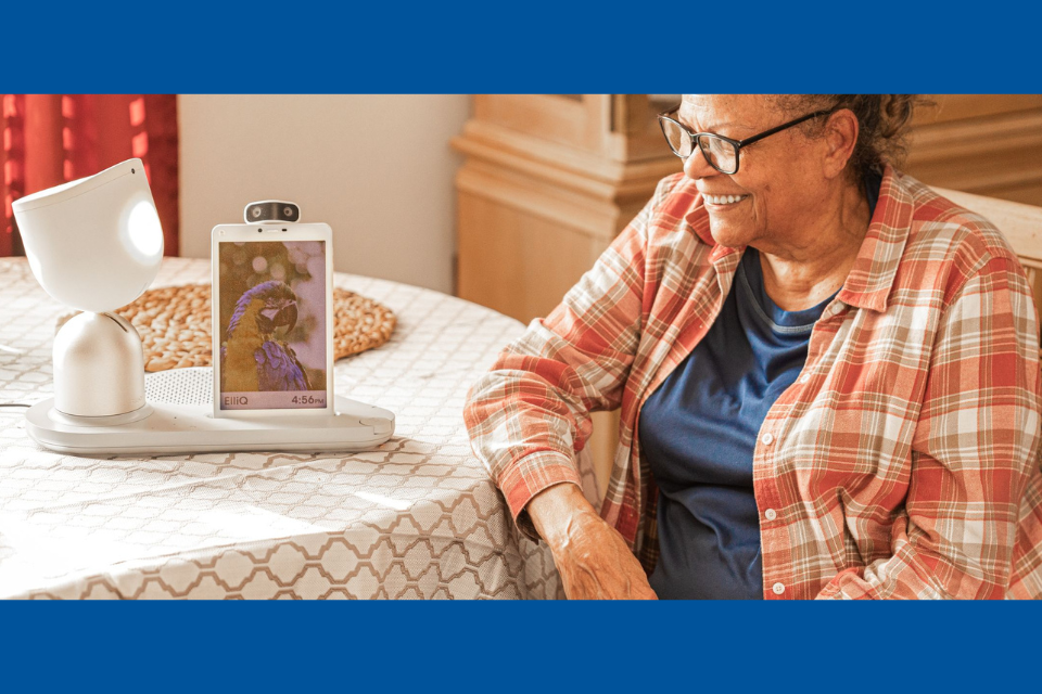 Older adult woman sitting at table looking at tabletop robot with amusement.