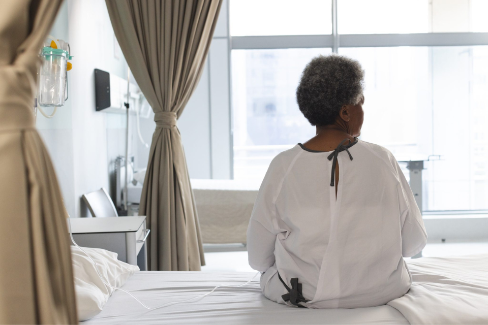Woman in hospital gown sitting on hospital bed, looking out window. Photo taken from behind woman.