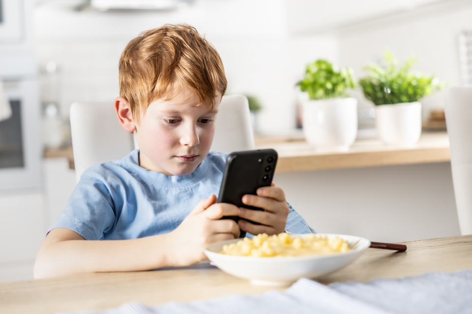 Child with bowl of mac & cheese; distracted by cell phone; not eating