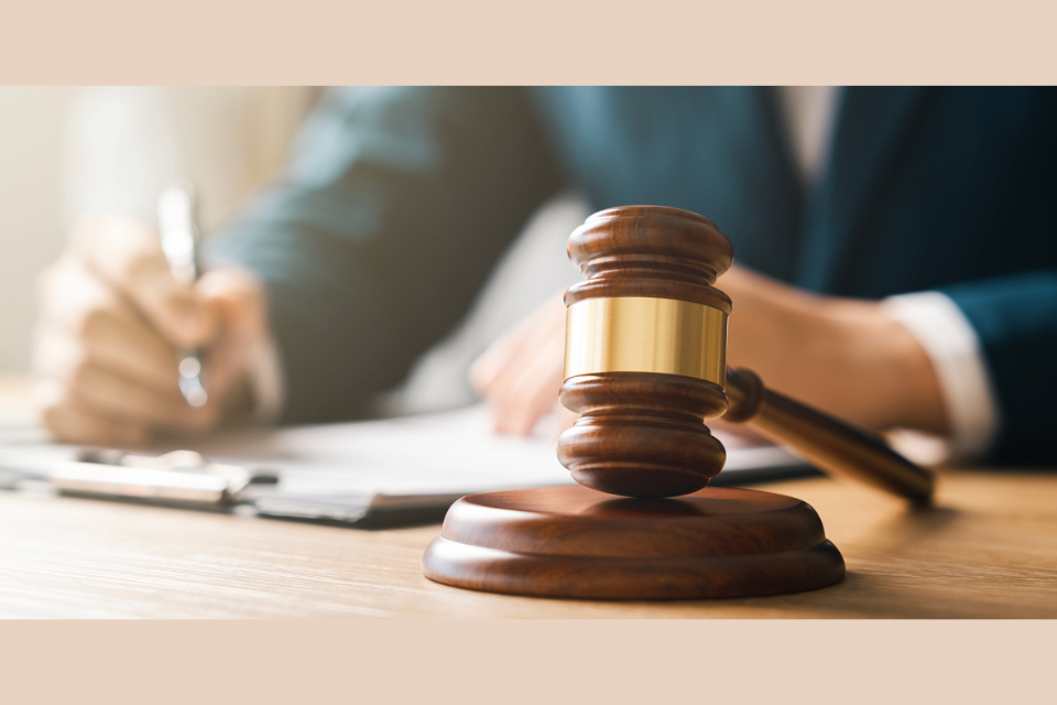 A wooden gavel with a brass band is placed on a wooden surface in the foreground. In the background, a person in a suit is holding a pen and writing on a clipboard or document. The image suggests a legal or judicial setting.