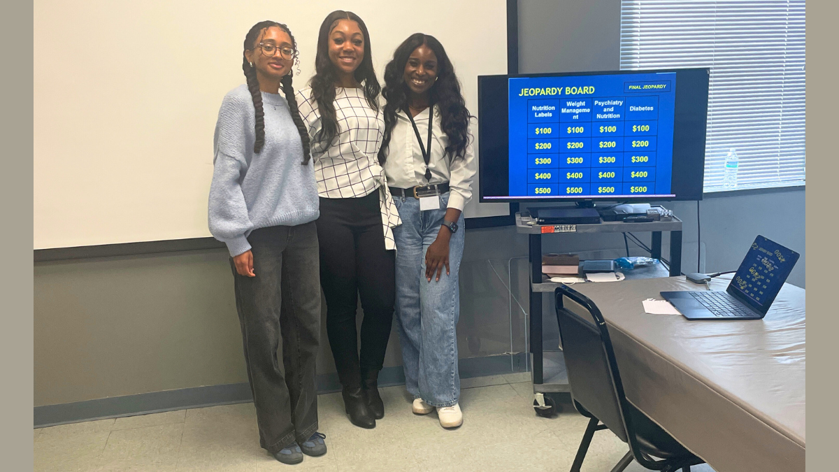 Three women standing beside a computer display with a "Jeopardy"-inspired board on the screen.