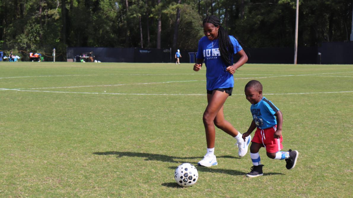 Duke Women's Soccer player dribbles the ball with young boy