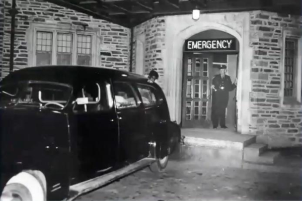Black and white image of old black car parked in front of Duke Emergency Department. Man standing in ED doorway.