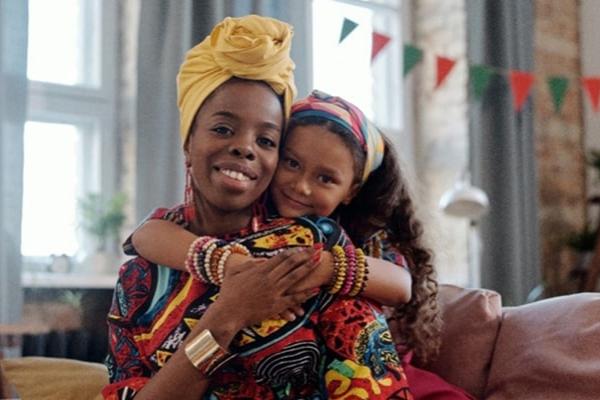 Black mother sitting down while daughter sits behind her and hands wrapped around her mothers chest 