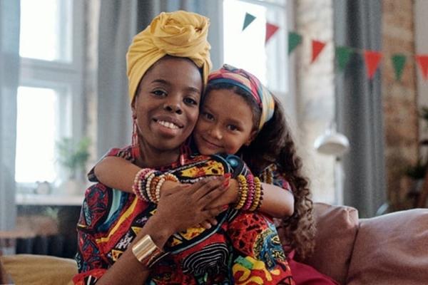 Black mother sitting down while daughter sits behind her and hands wrapped around her mothers chest