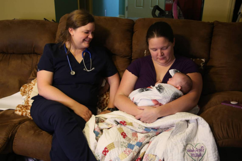 New mom holding baby in home with nurse looking on and smiling