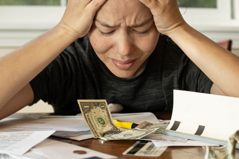 Person sitting at table with money and bills on table with hands holding head, looking distressed