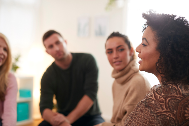 Three people talking in a group therapy setting. Fourth person on far left is cut off.