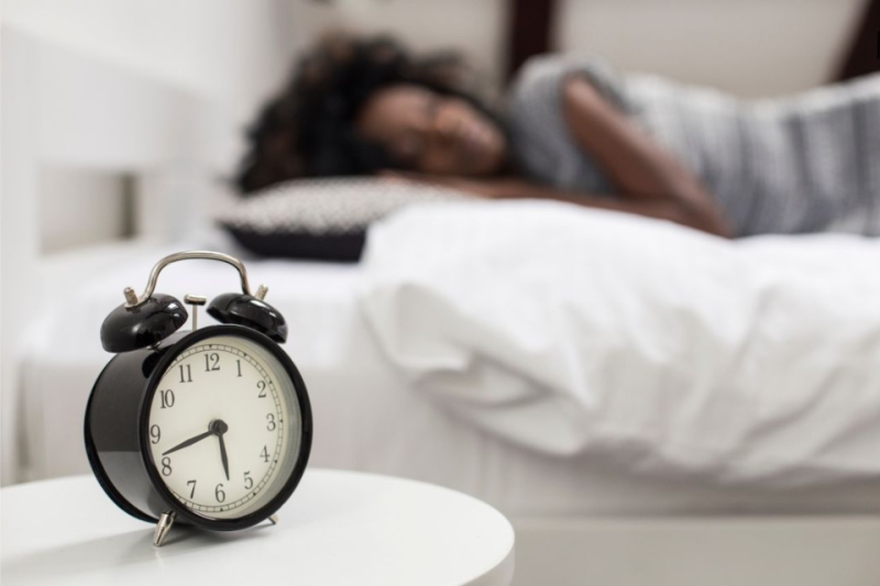 Alarm clock in foreground; woman sleeping on white sheets in background