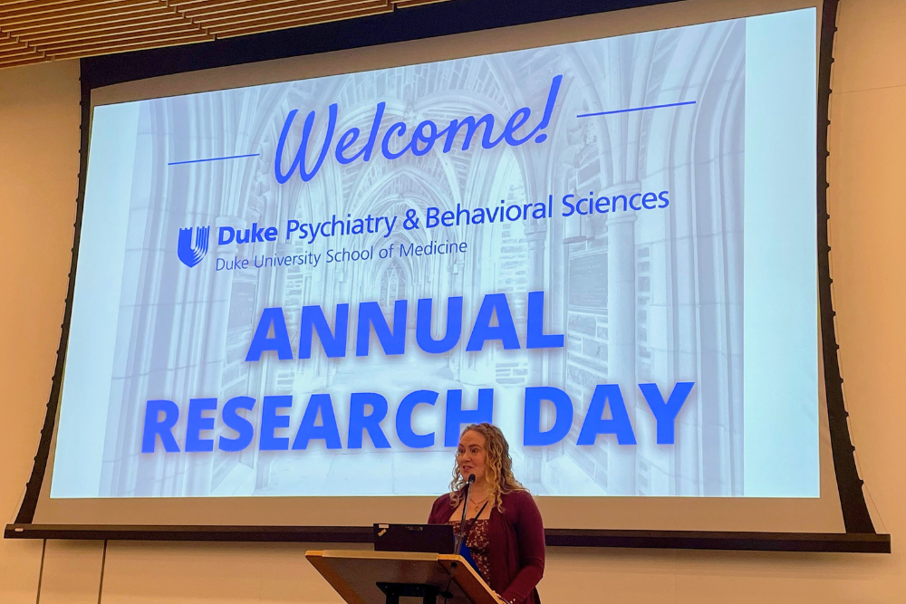 Woman standing at podium in front of projected slide, "Welcome Psychiatry & Behavioral Sciences Annual Research Day"