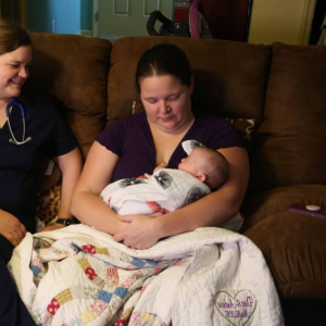 New mom holding baby in home with nurse looking on and smiling