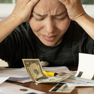Person sitting at table with money and bills on table with hands holding head, looking distressed