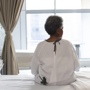 Woman in hospital gown sitting on hospital bed, looking out window. Photo taken from behind woman.