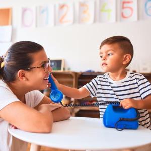 Young boy holds play phone to woman's ear in what appears to be a classroom or therapy setting.