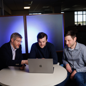 Jonathan Posner, Elliot Hill, and Matthew Engelhard look at a laptop together in a dark conference room..