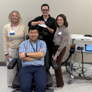One person sitting in TMS treatment chair with three other people gathered around the chair. TMS equipment to side.