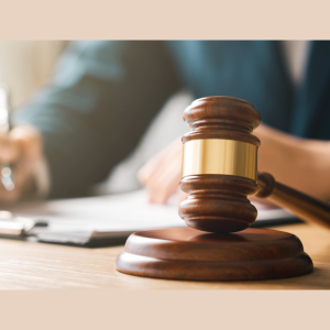 A wooden gavel with a brass band is placed on a wooden surface in the foreground. In the background, a person in a suit is holding a pen and writing on a clipboard or document. The image suggests a legal or judicial setting.