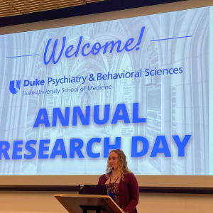 Woman standing at podium in front of projected slide, "Welcome Psychiatry & Behavioral Sciences Annual Research Day"